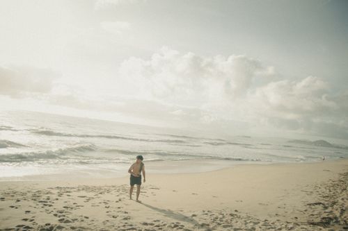 Free A child walks along a serene sandy beach with soft waves and overcast sky, evoking tranquility. Stock Photo
