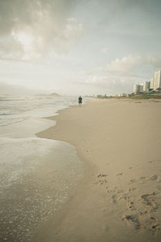 A lone person walks along a peaceful beach at daytime, beside city buildings.