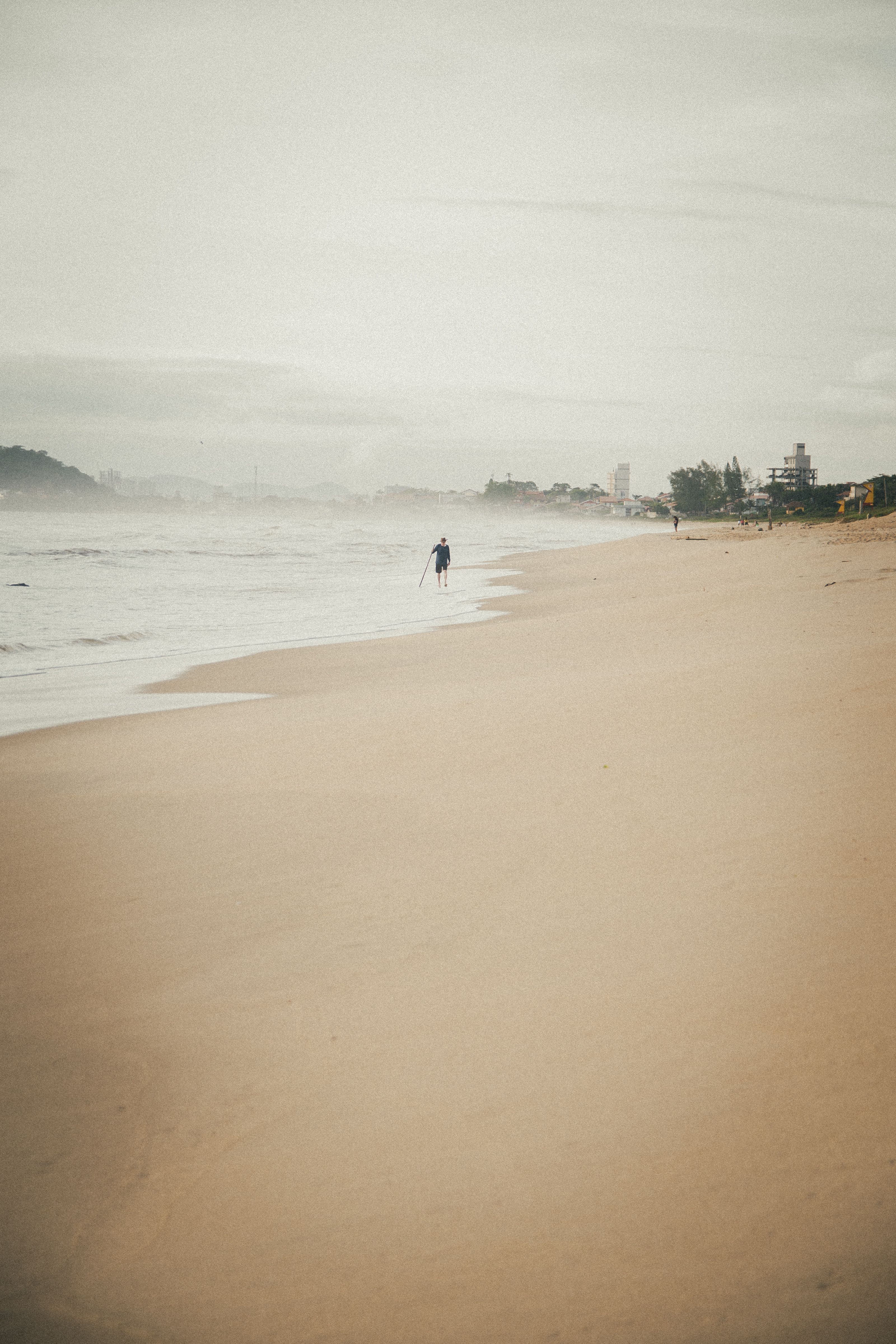 A lone figure walks along a misty, deserted beach under a cloudy sky, evoking a sense of solitude.