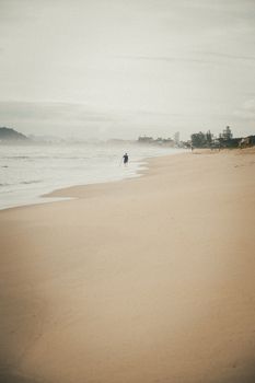 A lone figure walks along a misty, deserted beach under a cloudy sky, evoking a sense of solitude.