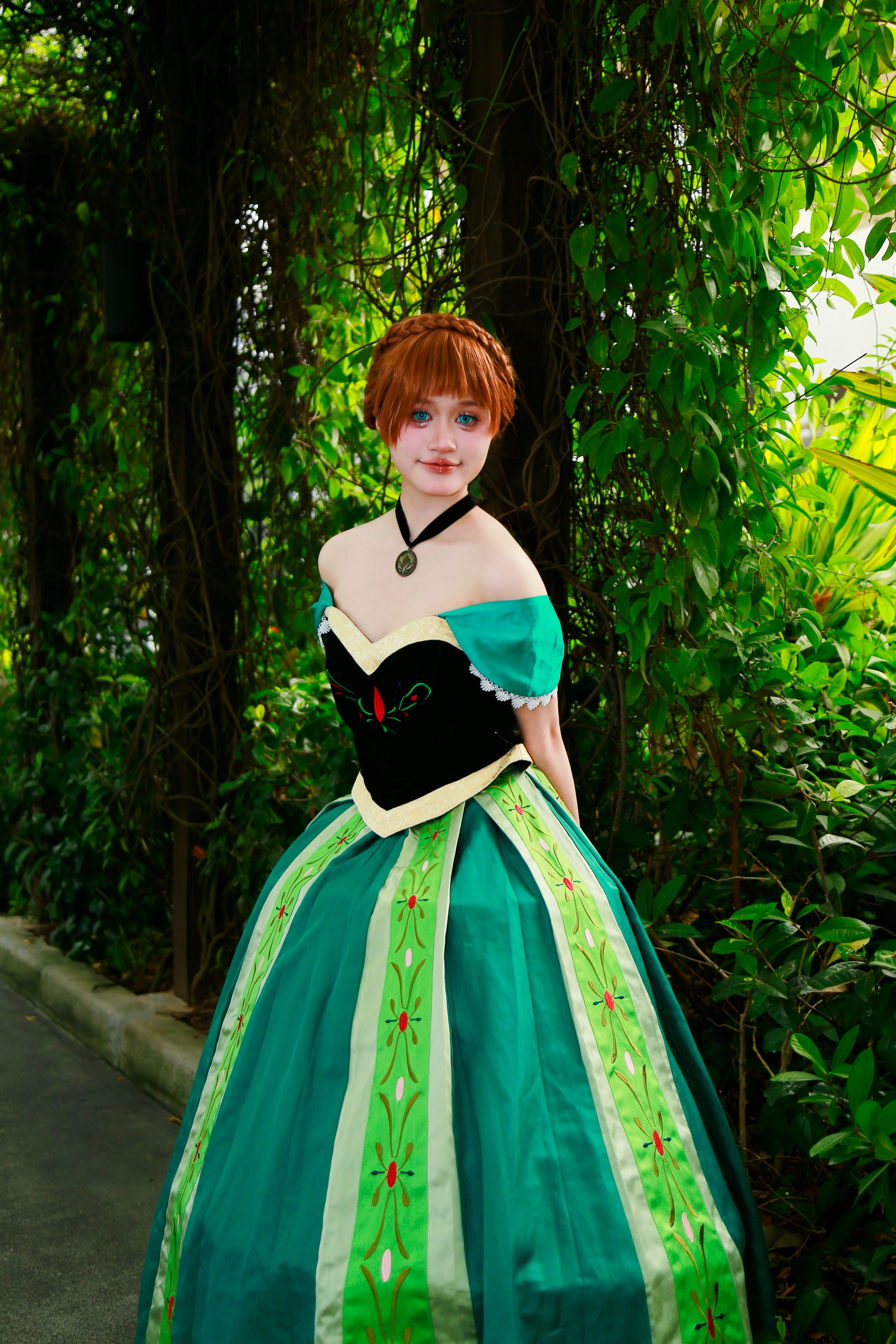 A woman in a green cosplay dress poses in a lush garden area outdoors.