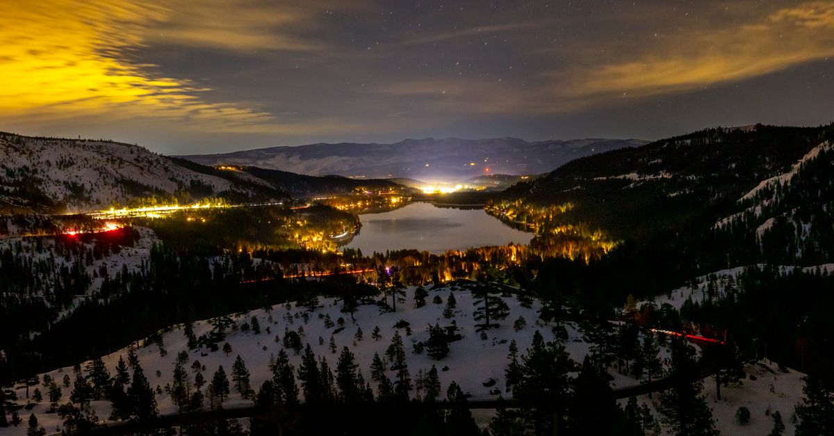 An enchanting night view of a starry sky over a snow-covered mountain lake, glowing with distant town lights.