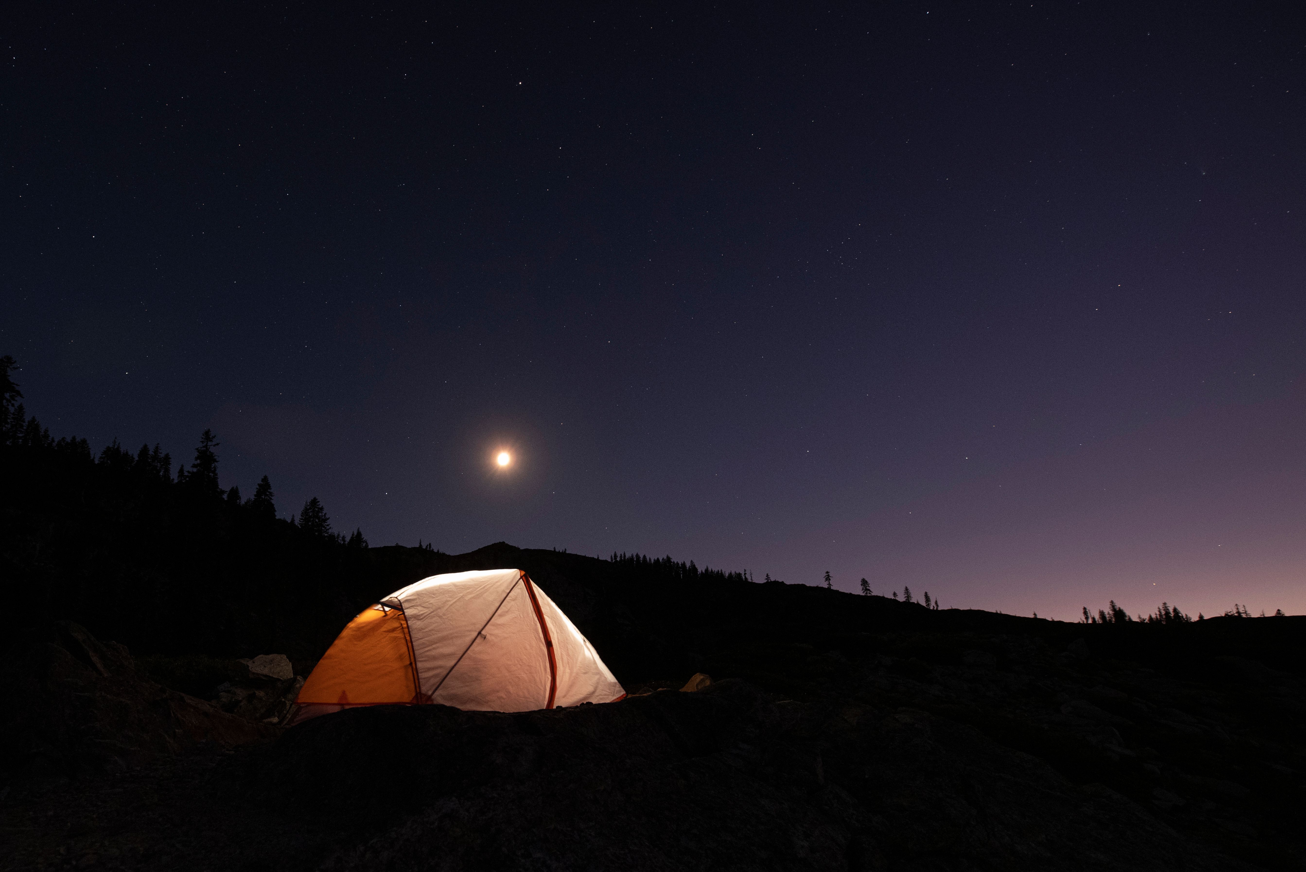 A serene night camping scene with a glowing tent under a star-filled sky and crescent moon.