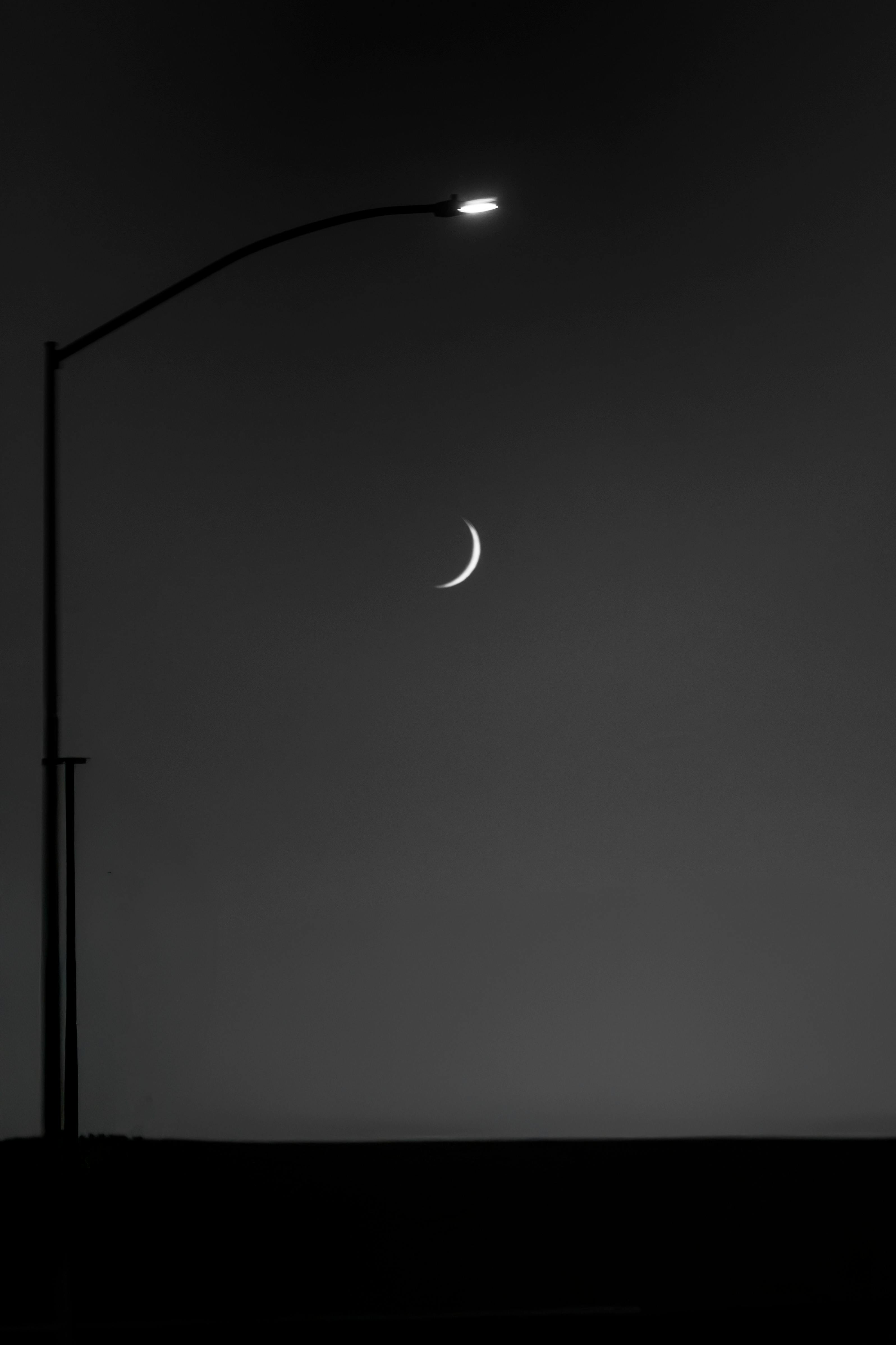grátis Fotografia melancólica em preto e branco, mostrando um poste de luz e uma lua crescente contra um céu noturno escuro. Foto profissional