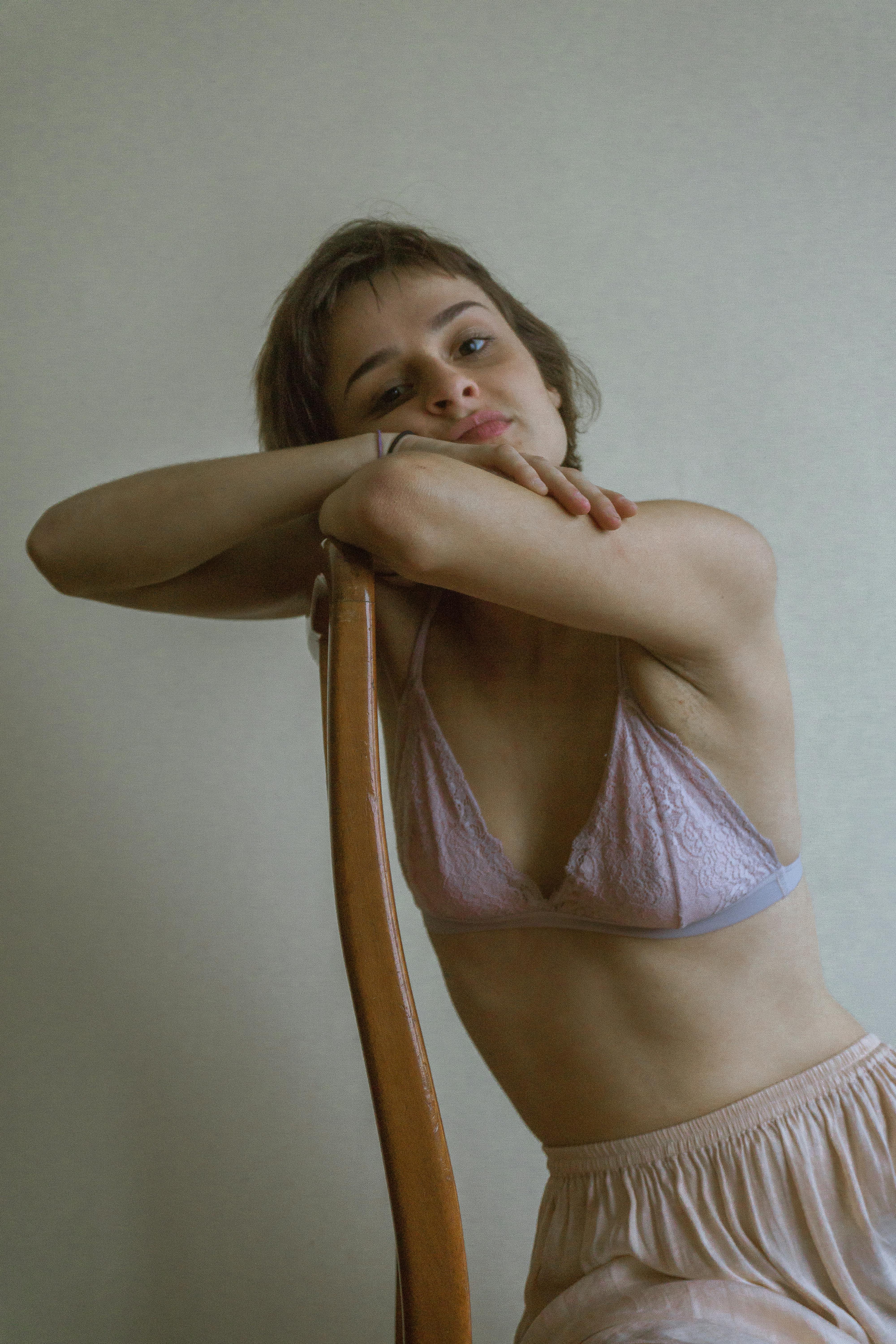 Bored young female in bra and skirt sitting on chair and resting head on hands against white wall and looking at camera