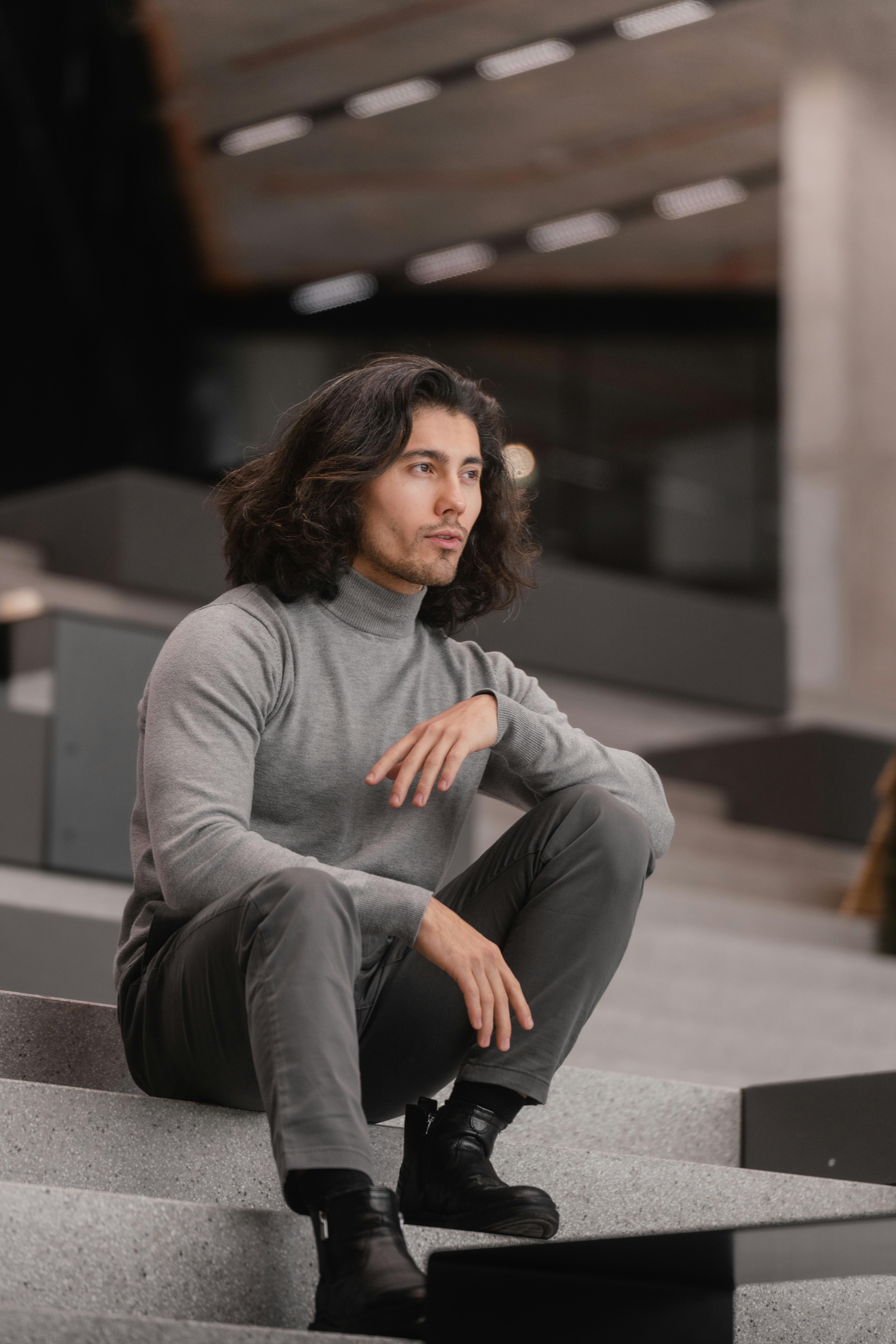 Zadarmo Mladý muž s dlhými vlasmi v roláku sediaci na schodoch, odrážajúci modernú módu. Fotka z fotobanky