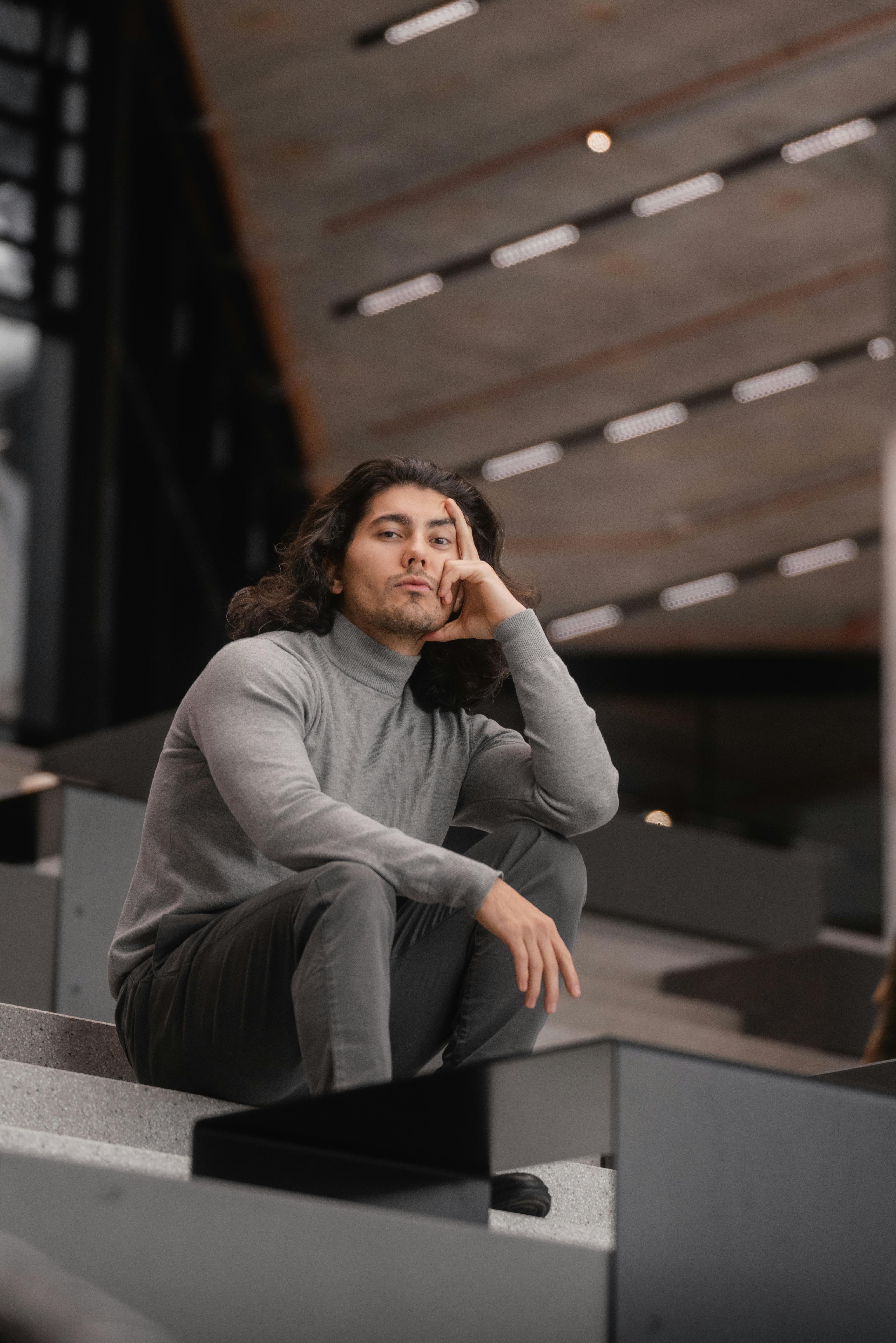 Gratis Un uomo con un maglione grigio seduto su una scala moderna, che fonde lo stile urbano con l'architettura. Foto a disposizione