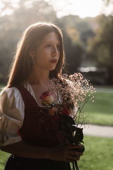 A serene portrait of a young woman in traditional Austrian attire holding a flower bouquet in a park.