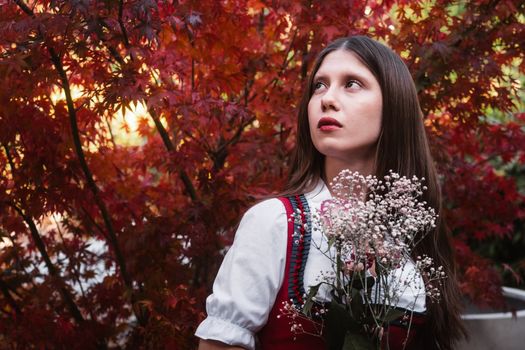 A woman in traditional Austrian dress stands with flowers, framed by vibrant autumn leaves.