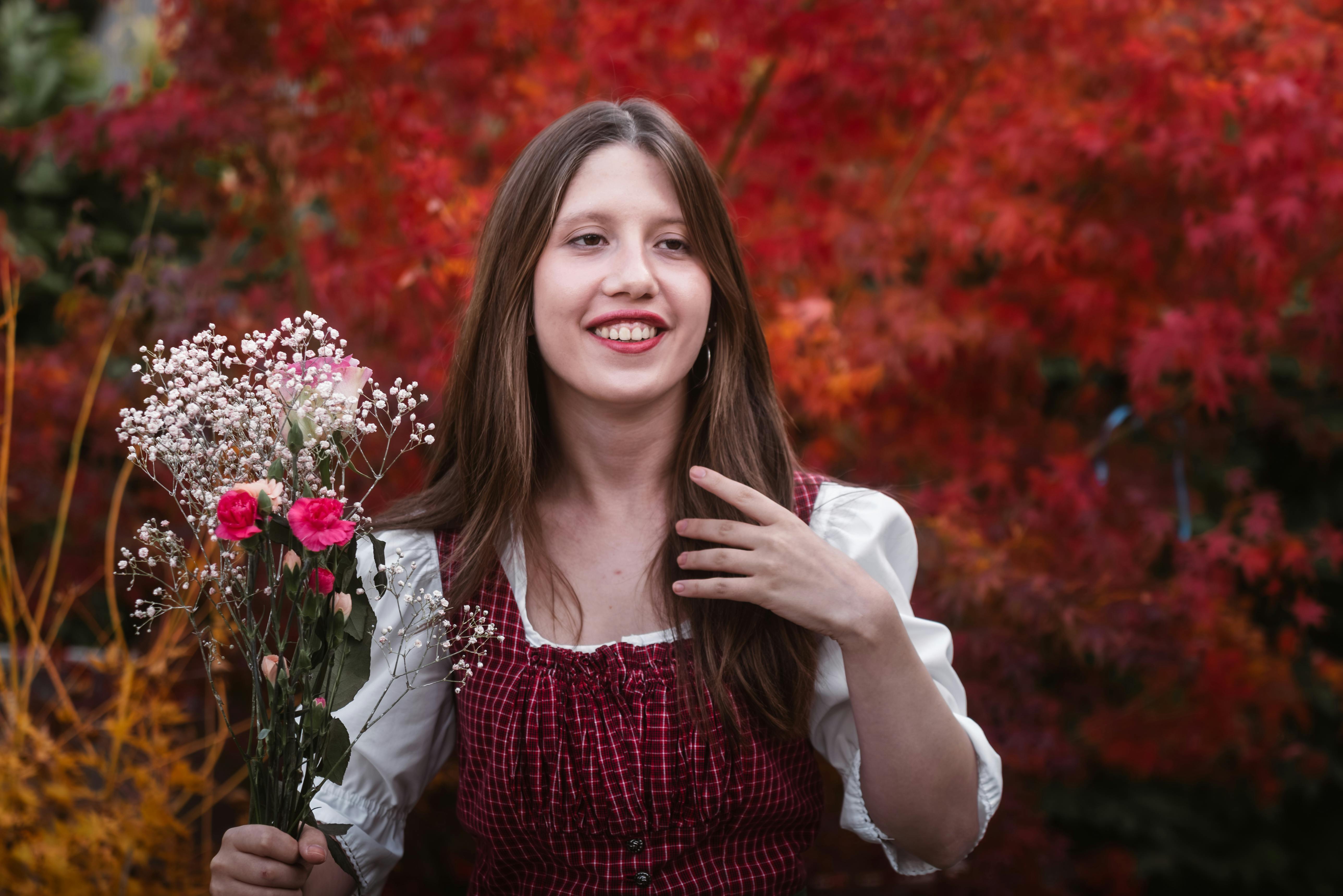 Gratis Una giovane donna in abito tradizionale tiene in mano dei fiori in mezzo al vivace fogliame autunnale a Graz, in Austria. Foto a disposizione