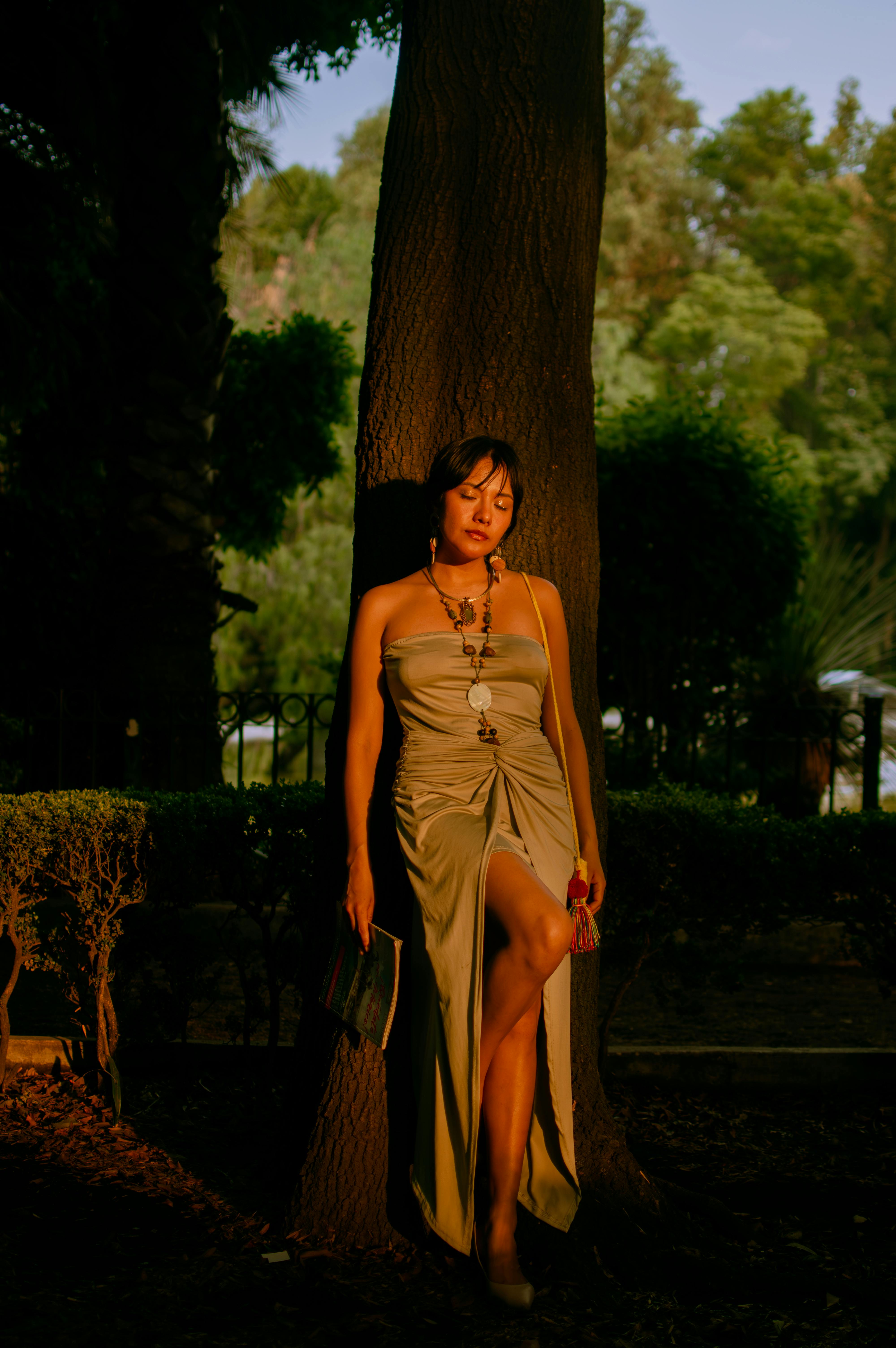 Free Portrait of a woman in a long dress leaning against a tree at sunset in a garden. Stock Photo