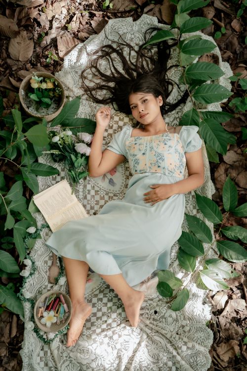 Free Young woman in a light dress reclines on a blanket surrounded by nature in a forest setting. Stock Photo