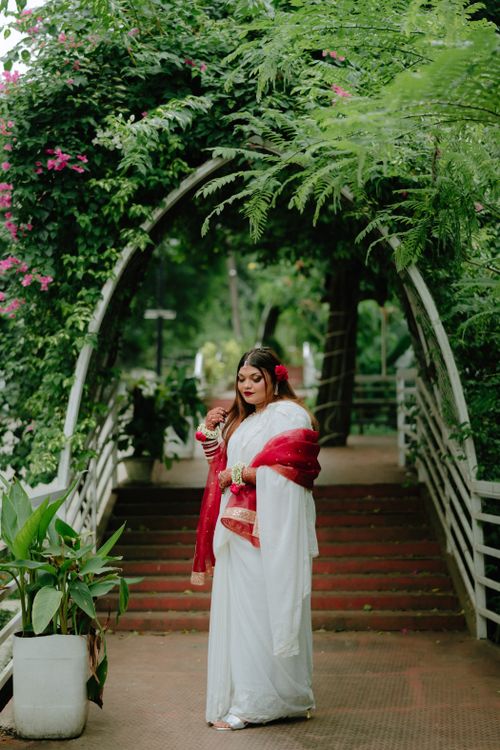 Free Elegant woman in white and red sari poses under lush greenery archway outdoors. Stock Photo