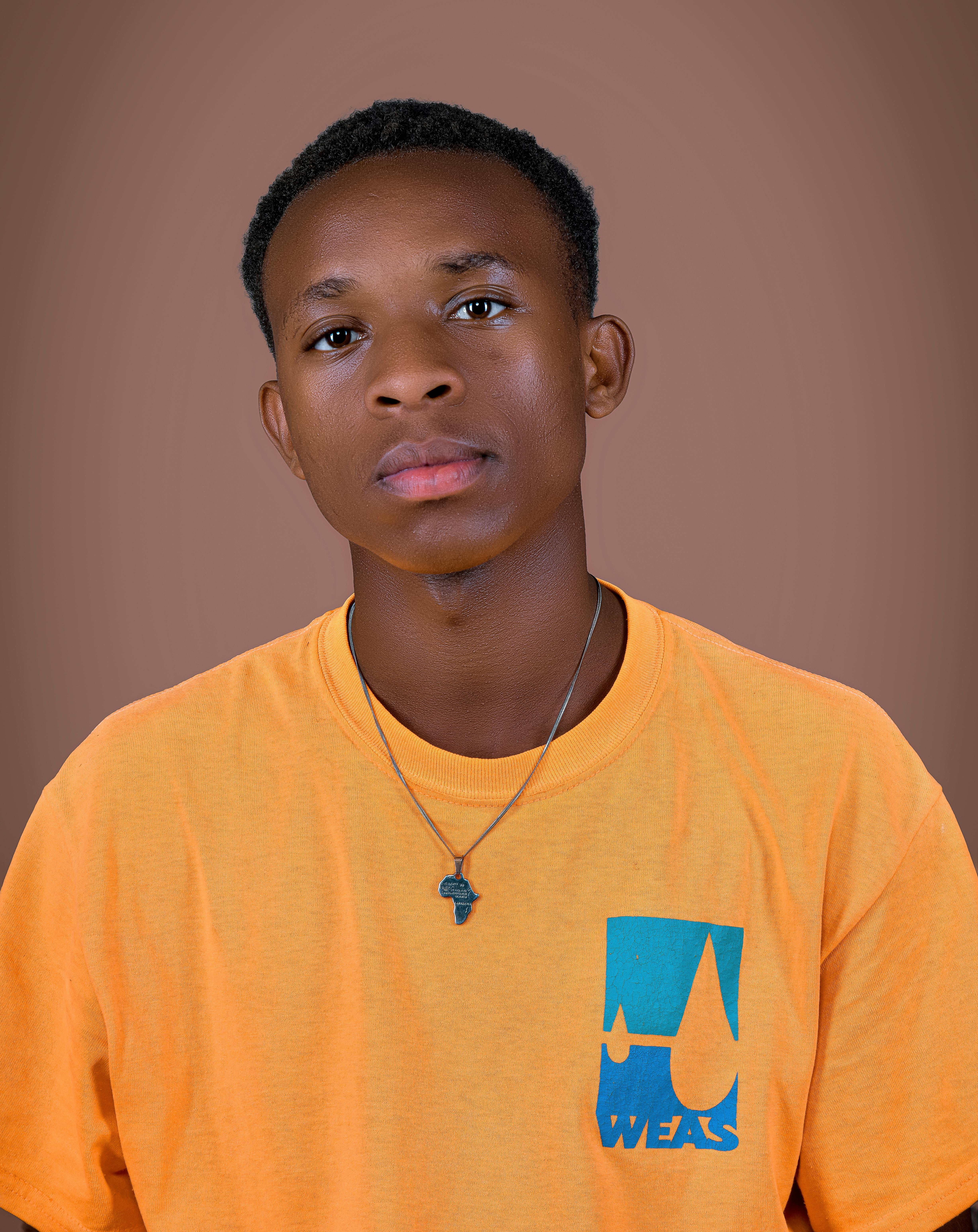 Portrait of a young man wearing an orange shirt and a pendant necklace against a brown backdrop.