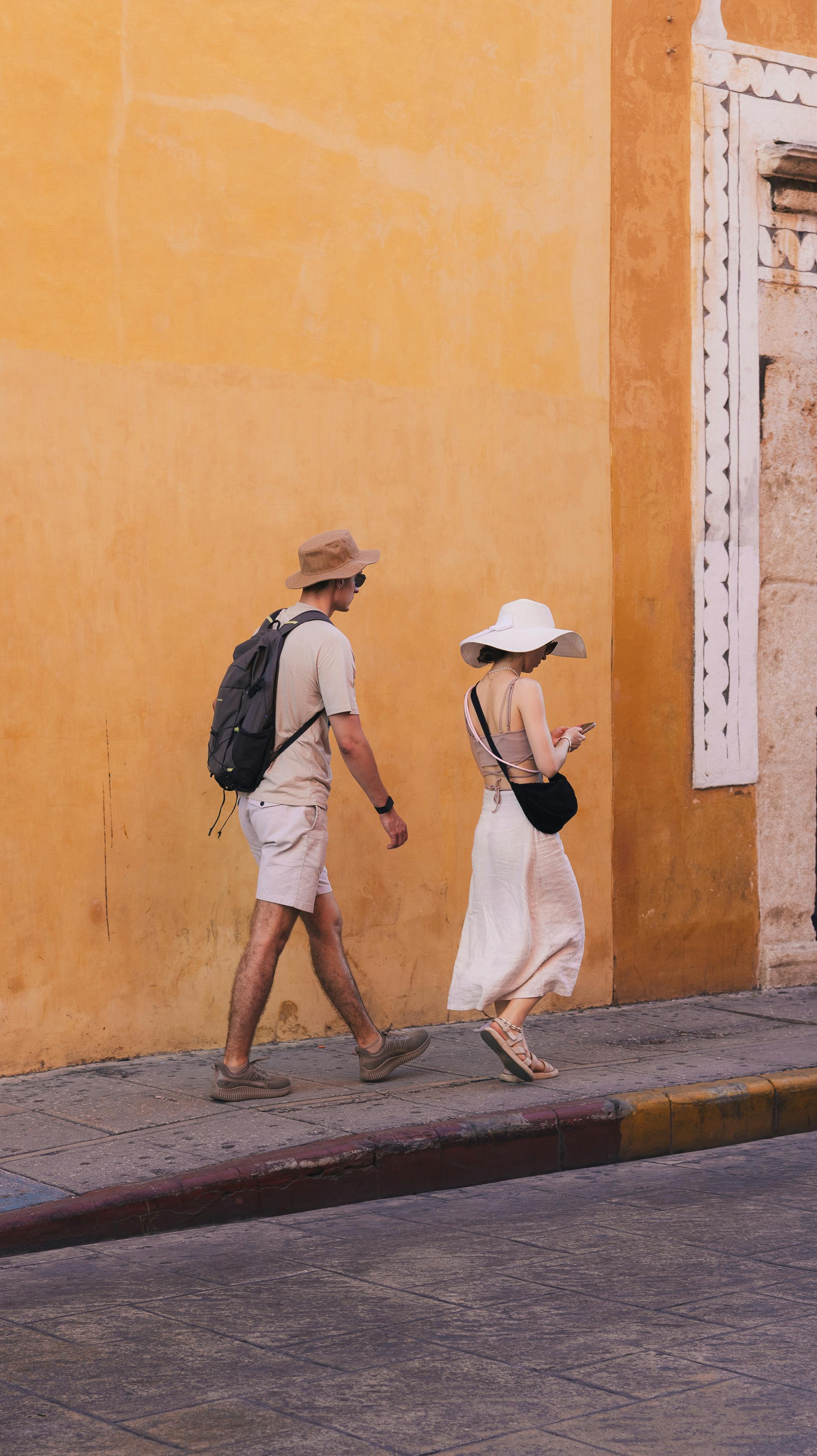 Free Couple strolling along a vibrant yellow wall, embracing summer with hats and light attire. Stock Photo