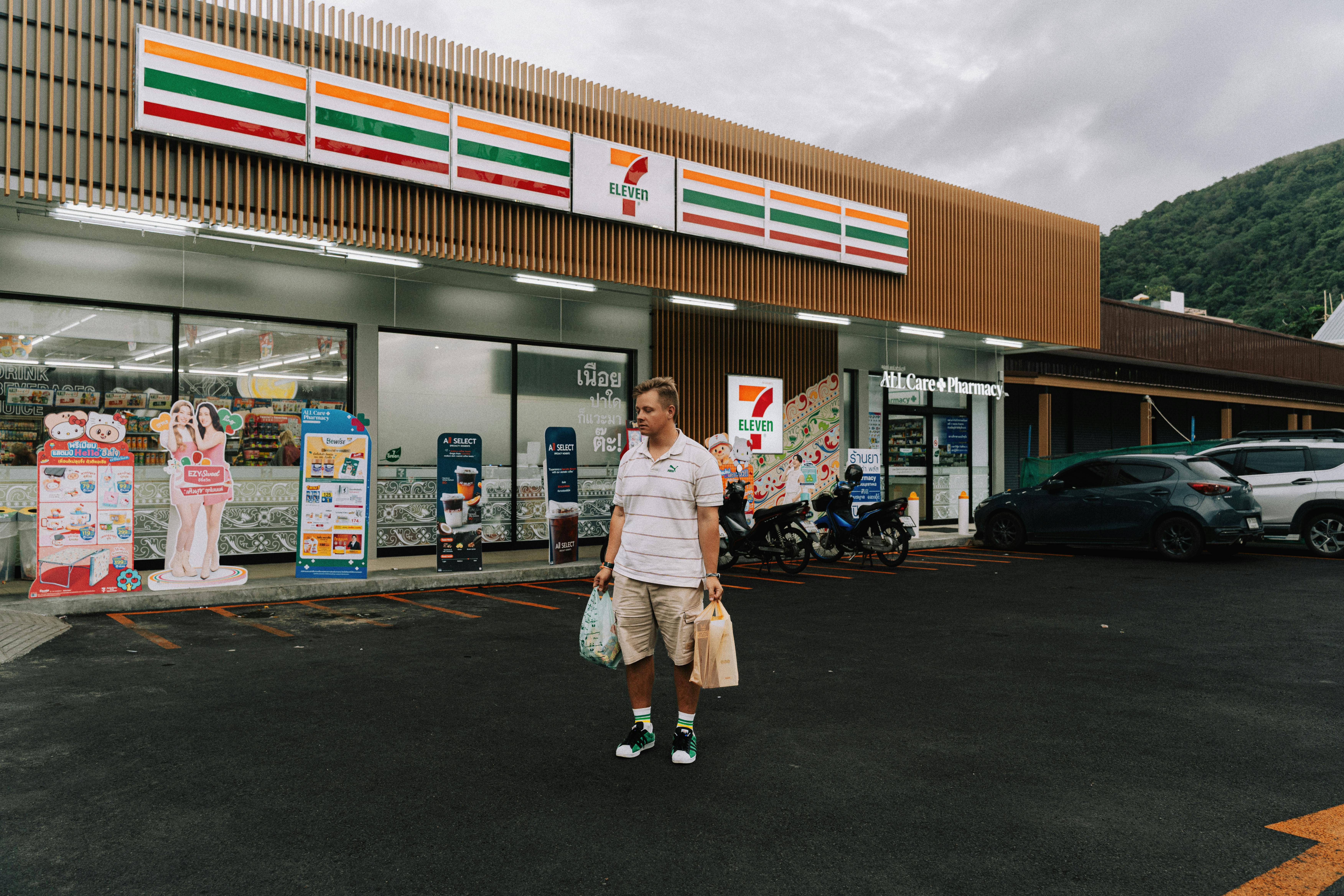 Man with shopping bags outside 7-Eleven in Phuket, showcasing a modern convenience store.