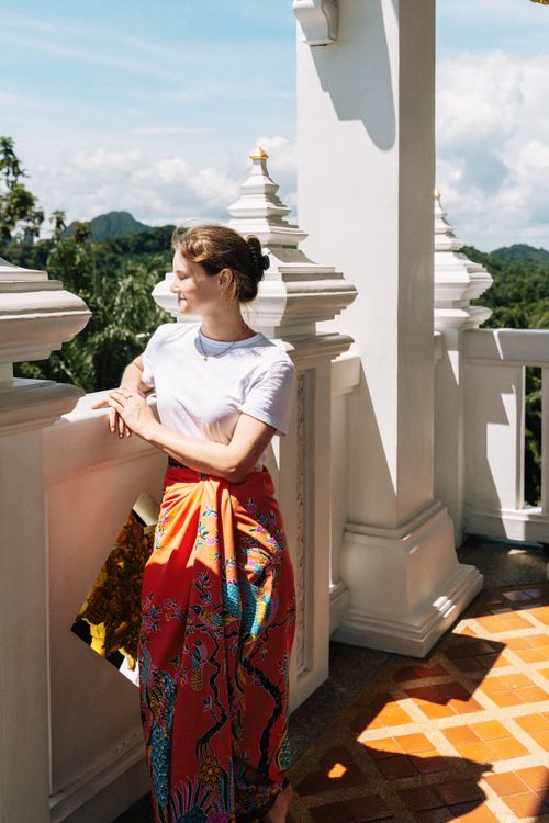 Free Woman in a vibrant skirt stands at a Krabi temple terrace, enjoying the sunny view. Stock Photo