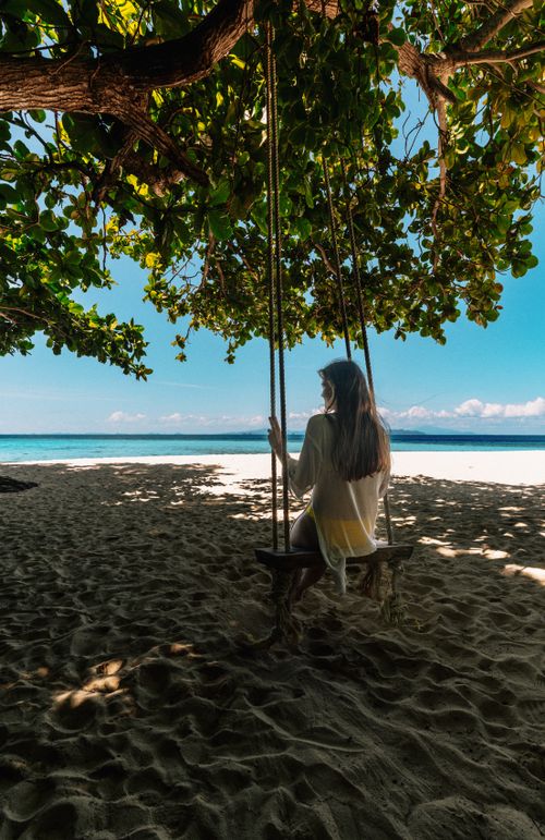Free Woman relaxing on a swing under a lush tree canopy by the serene beach in Ao Nang, Thailand. Stock Photo