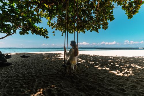 Free Serene tropical beach scene with a woman on a swing under lush trees. Stock Photo