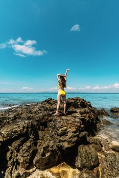 Woman in yellow bikini standing on coastal rocks, gazing at tranquil sea, under clear blue sky.