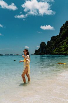 A woman in a yellow beach cover-up walks along the shore at Ko Panyi, Thailand, with clear blue water and lush cliffs.