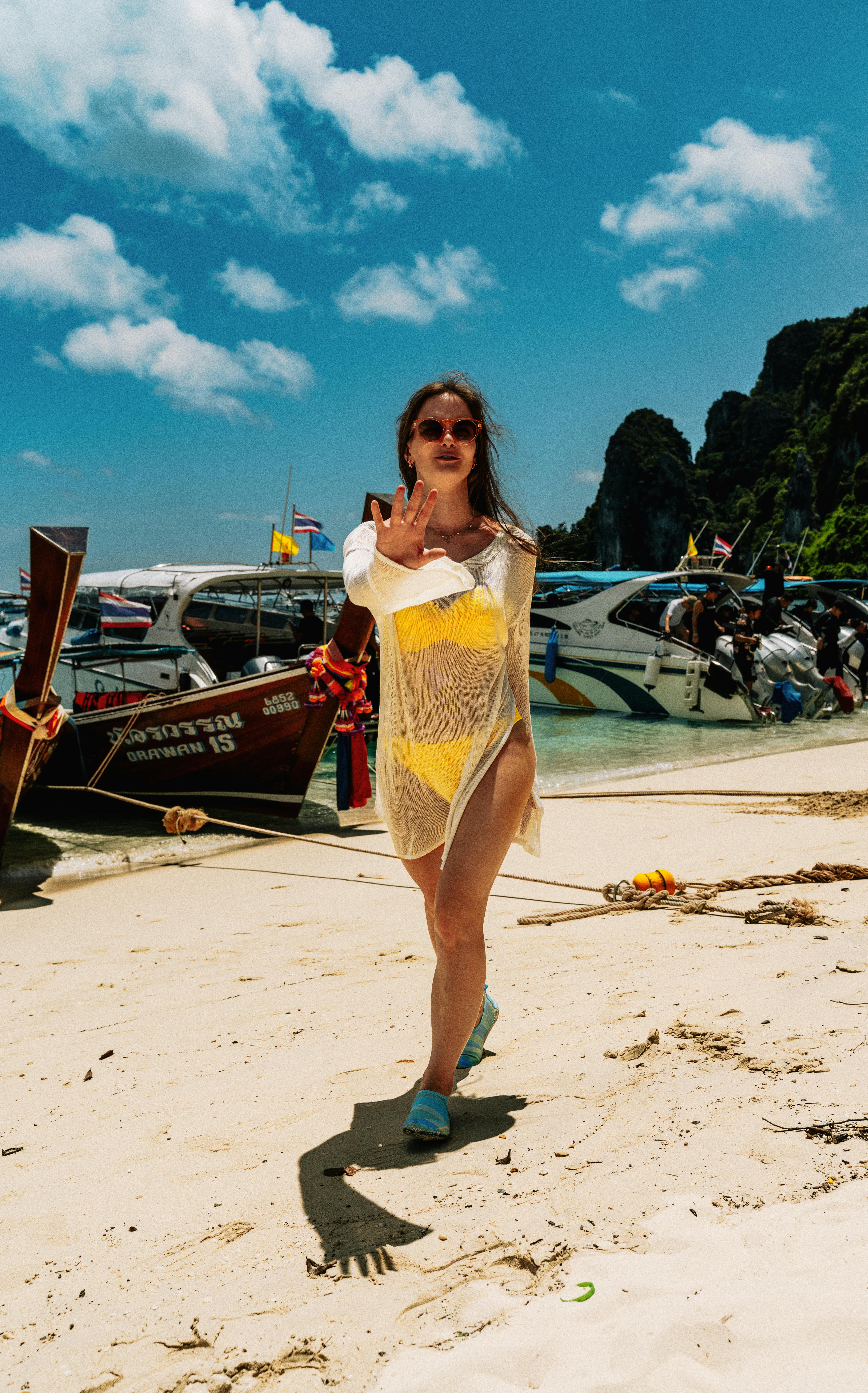 Stylish woman in a yellow bikini poses on the beaches of Ko Panyi, Thailand with boats and rocky cliffs in the background.