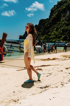 A young woman strolls on the sunny beach of Ko Panyi, Thailand, with boats and lush cliffs in the background.