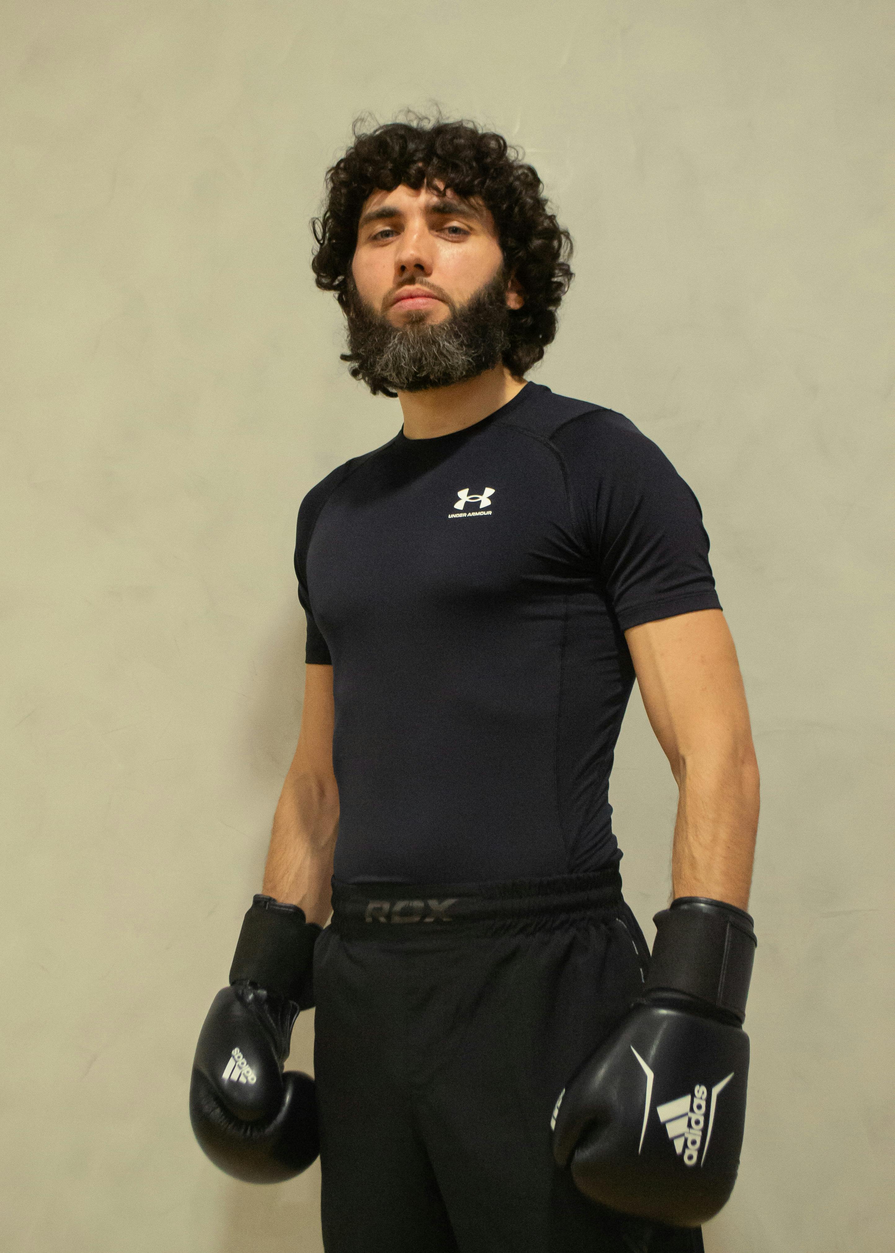 Bearded male boxer in black athletic wear poses confidently with boxing gloves in gym setting.