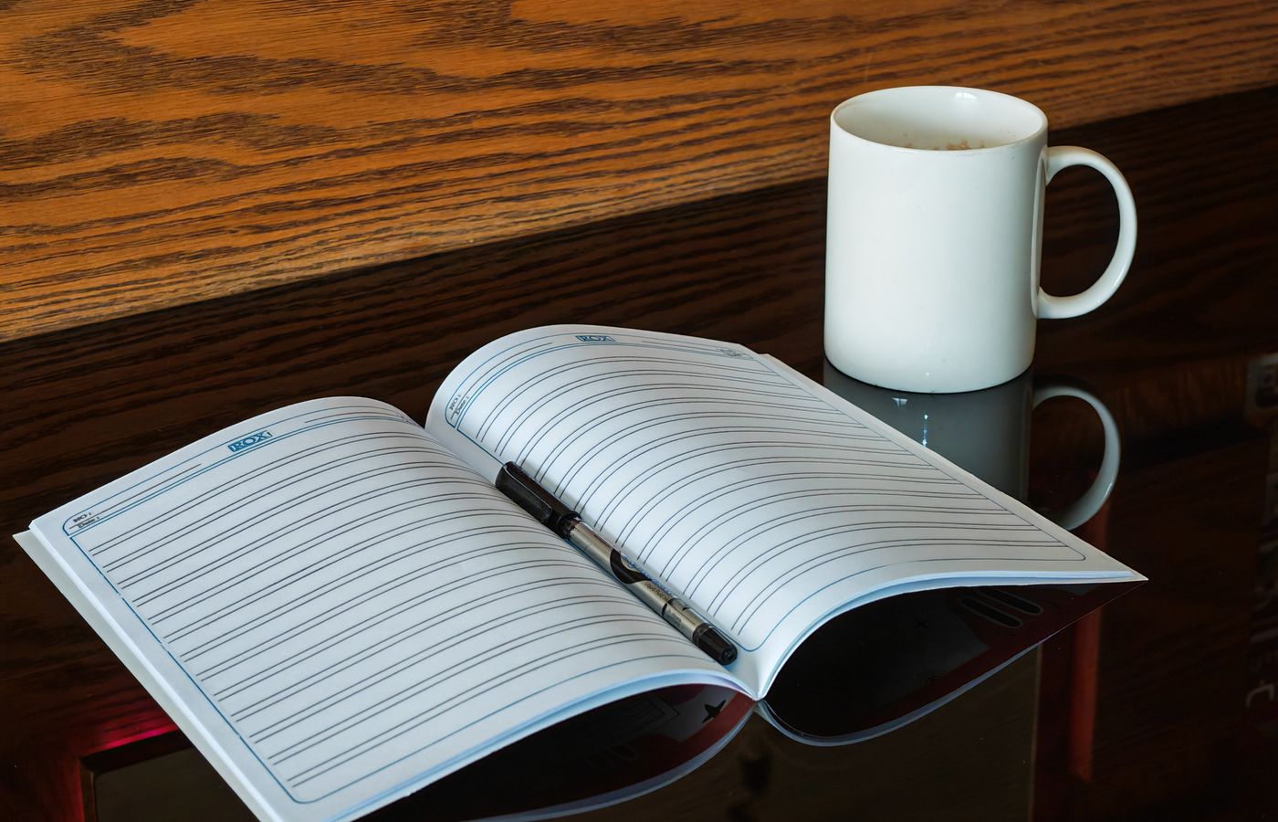 An open notebook with a pen beside a coffee mug on a reflective table.
