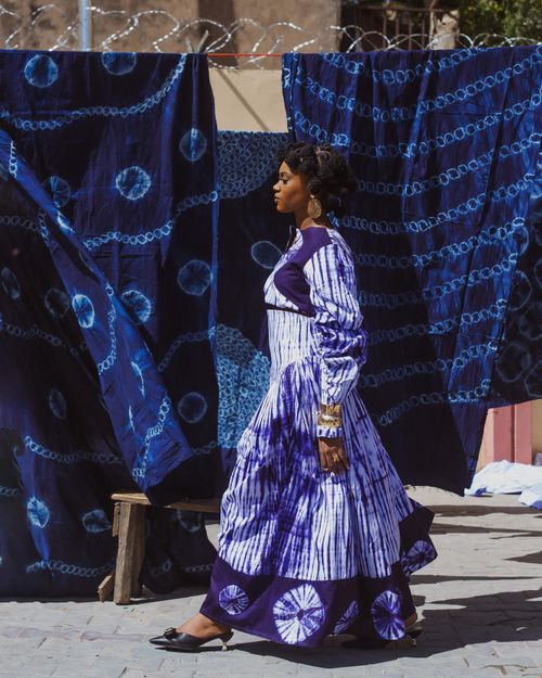 Free Confident woman in vibrant traditional dress walking past tie-dyed fabrics outdoors. Stock Photo