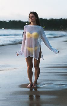 A woman standing on a beach in Phuket during twilight wearing a bikini under a transparent cover-up.