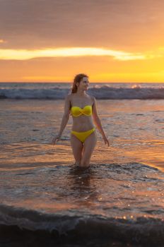 Adult woman in yellow bikini walking through ocean waves during a stunning sunset on a Phuket beach.