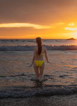 A woman in a yellow bikini stands in the ocean at sunset in Phuket, Thailand, embracing tranquillity.