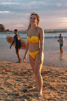 A woman in a yellow bikini stands on a sandy beach in Phuket, Thailand, during a beautiful sunset.