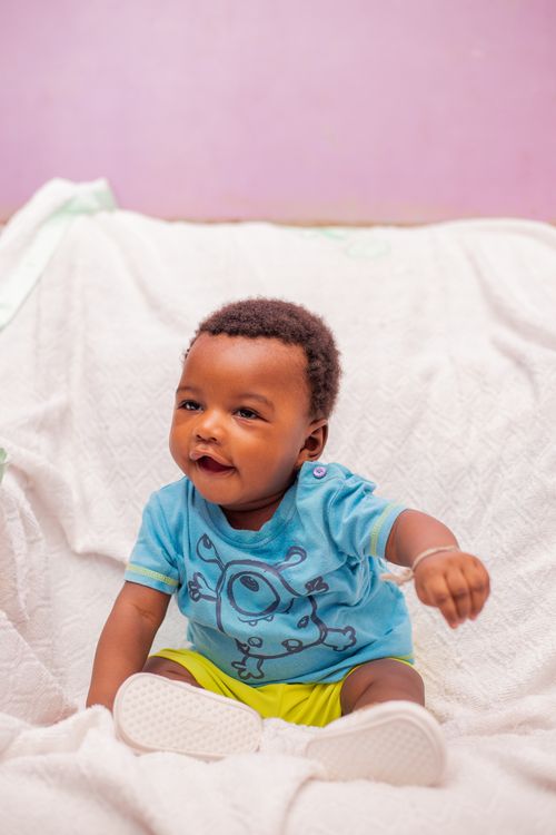 Free Cute baby wearing a blue shirt sitting indoors, smiling happily on a white blanket. Stock Photo