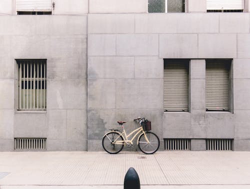 Bicycle with basket near handlebar parked against gray concrete building wall on paved sidewalk during daytime