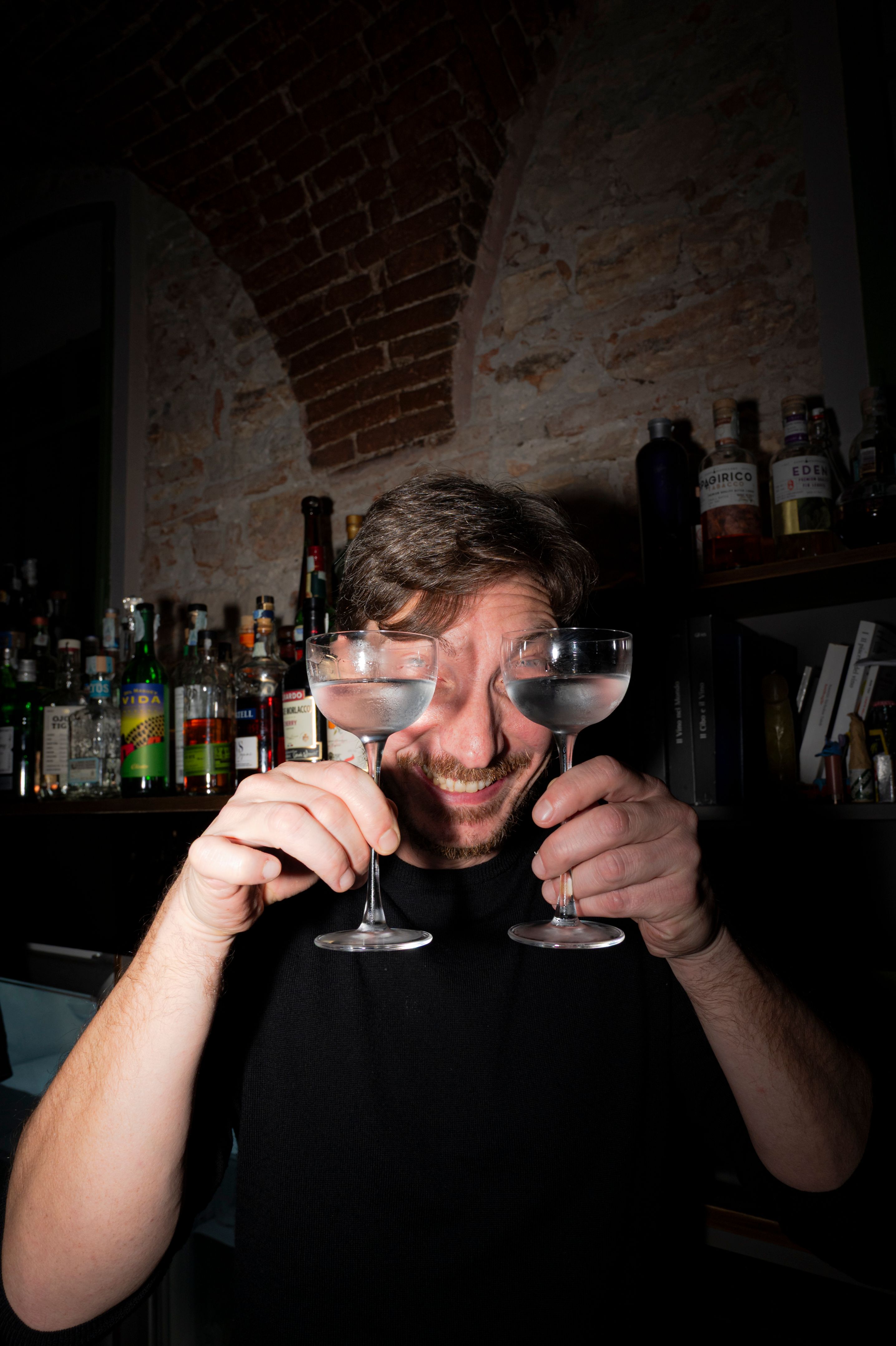Man happily holding cocktail glasses in a cozy bar in Brescia, Italy.