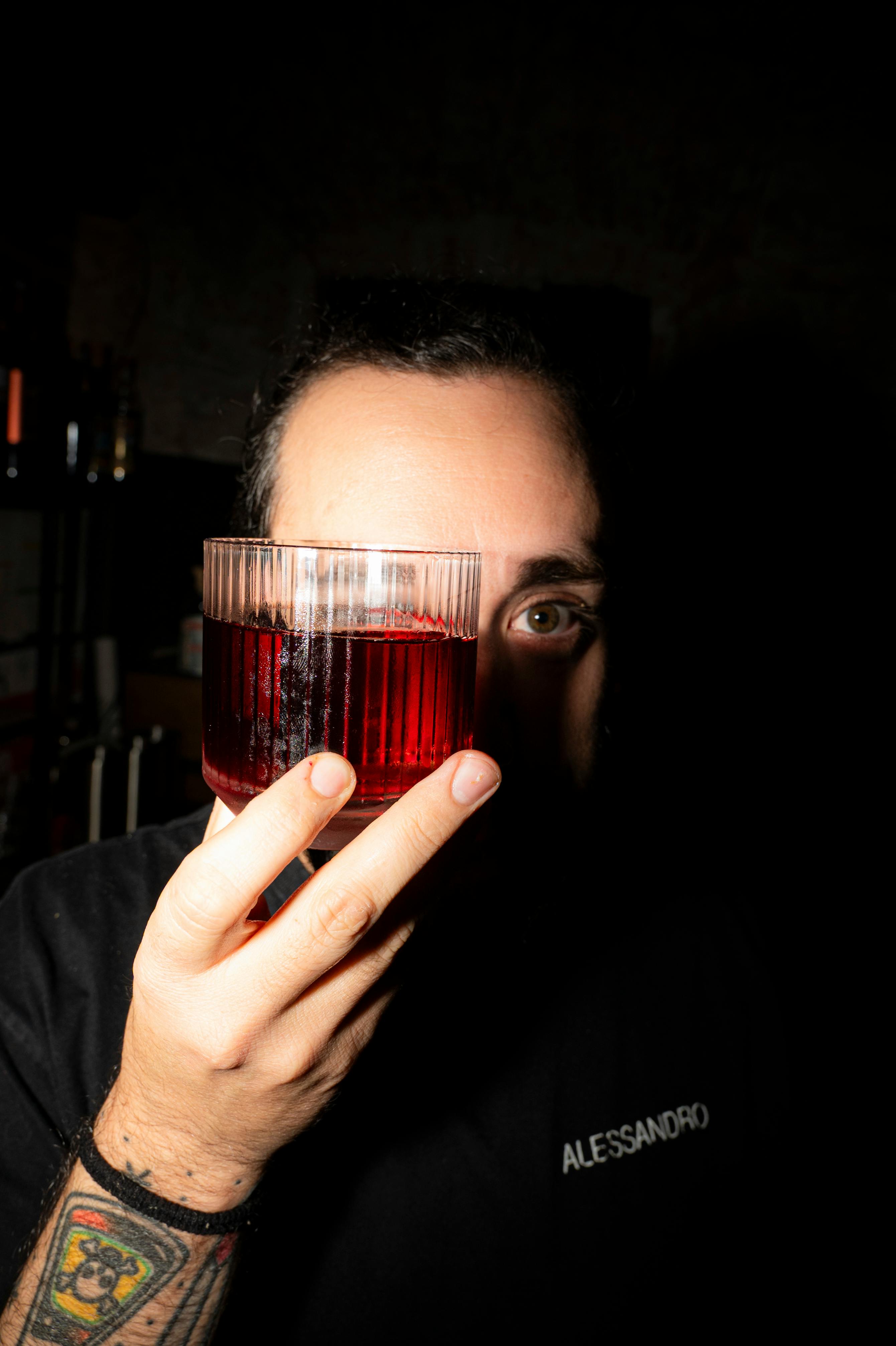 A man holds a glass of red drink covering part of his face in a dimly lit room.
