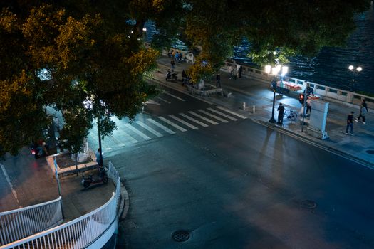 Aerial view of an illuminated city street and riverside at night with people walking.