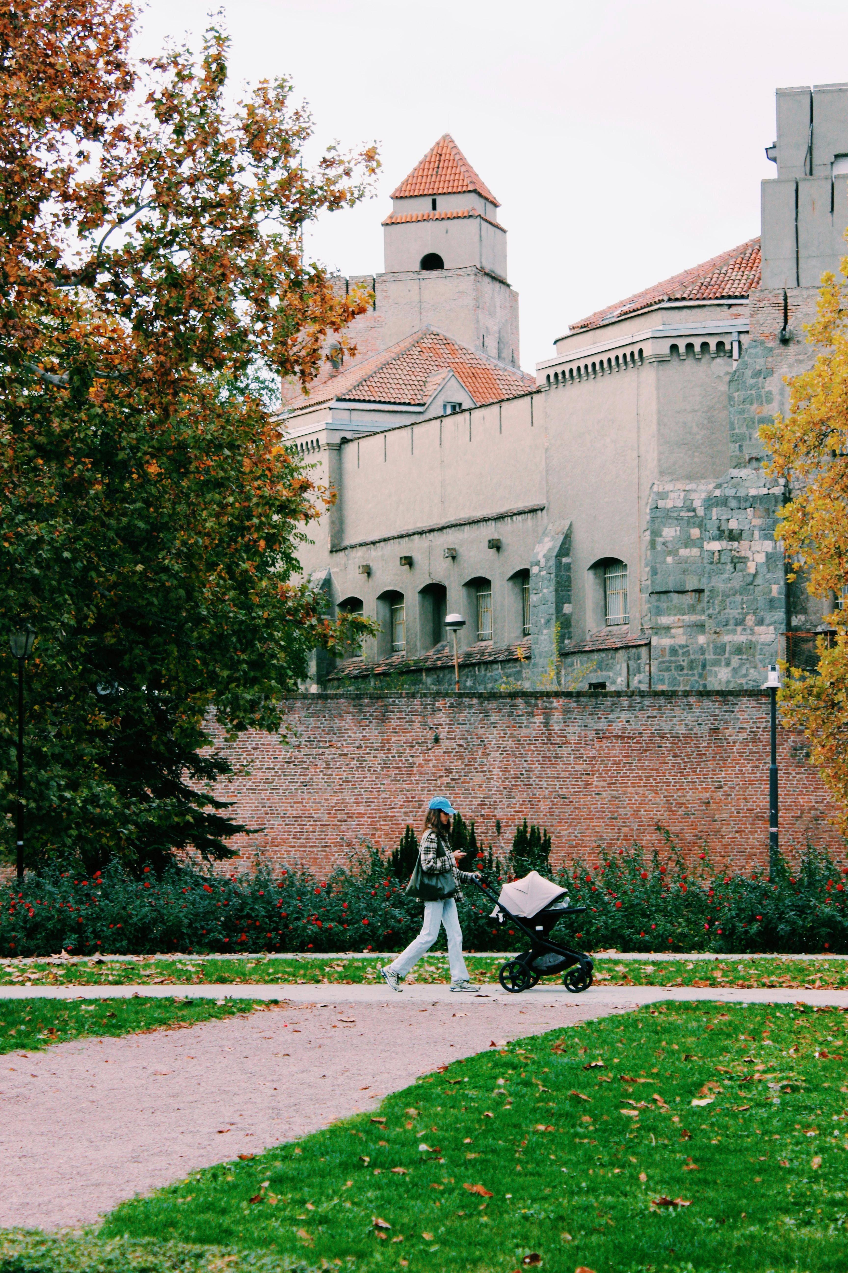 Woman strolls with baby stroller in autumn near historic city wall.