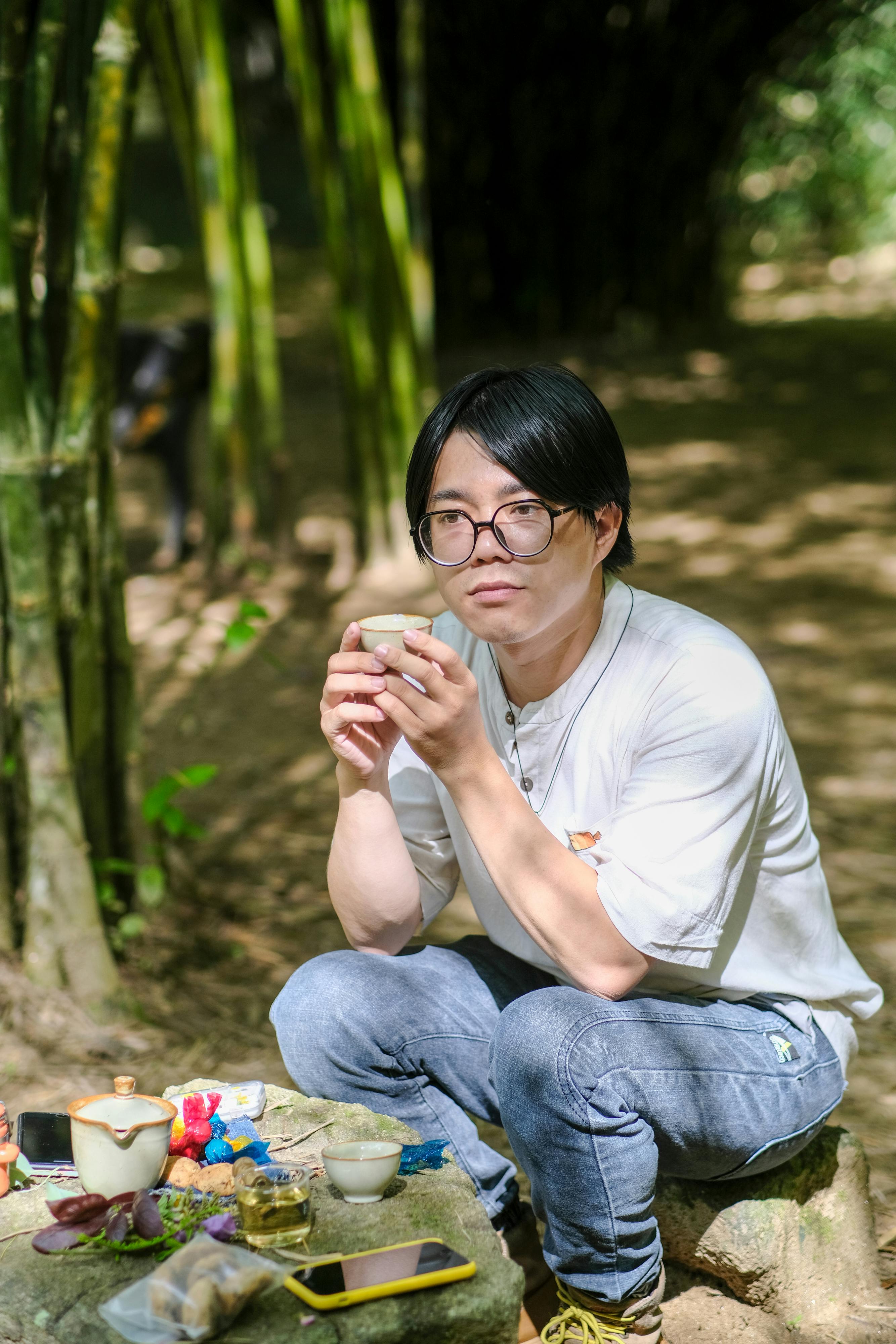 Man sitting outdoors in a bamboo grove, enjoying tea, wearing glasses.