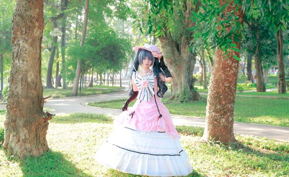 A cosplayer in a pink Victorian gown poses in a sunny park with lush greenery.
