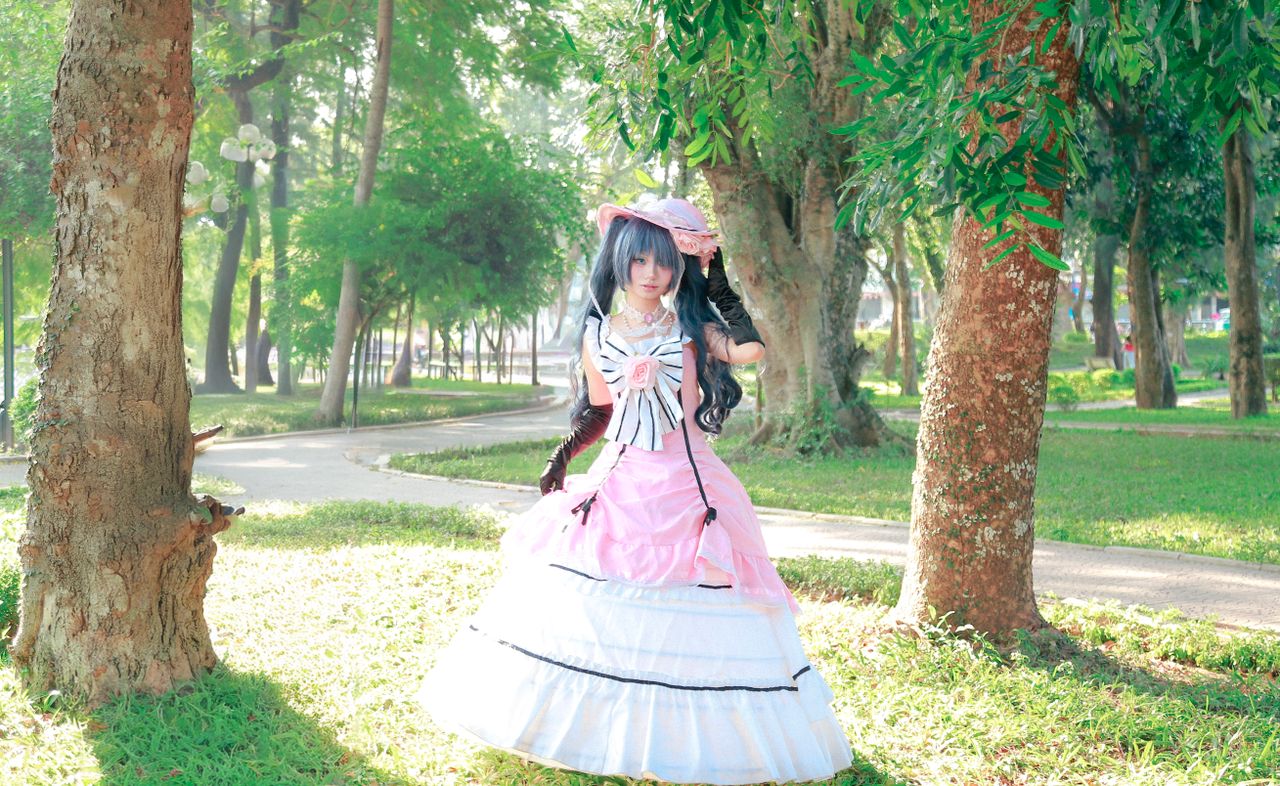 a cosplayer in a pink victorian gown poses in a sunny park with lush g