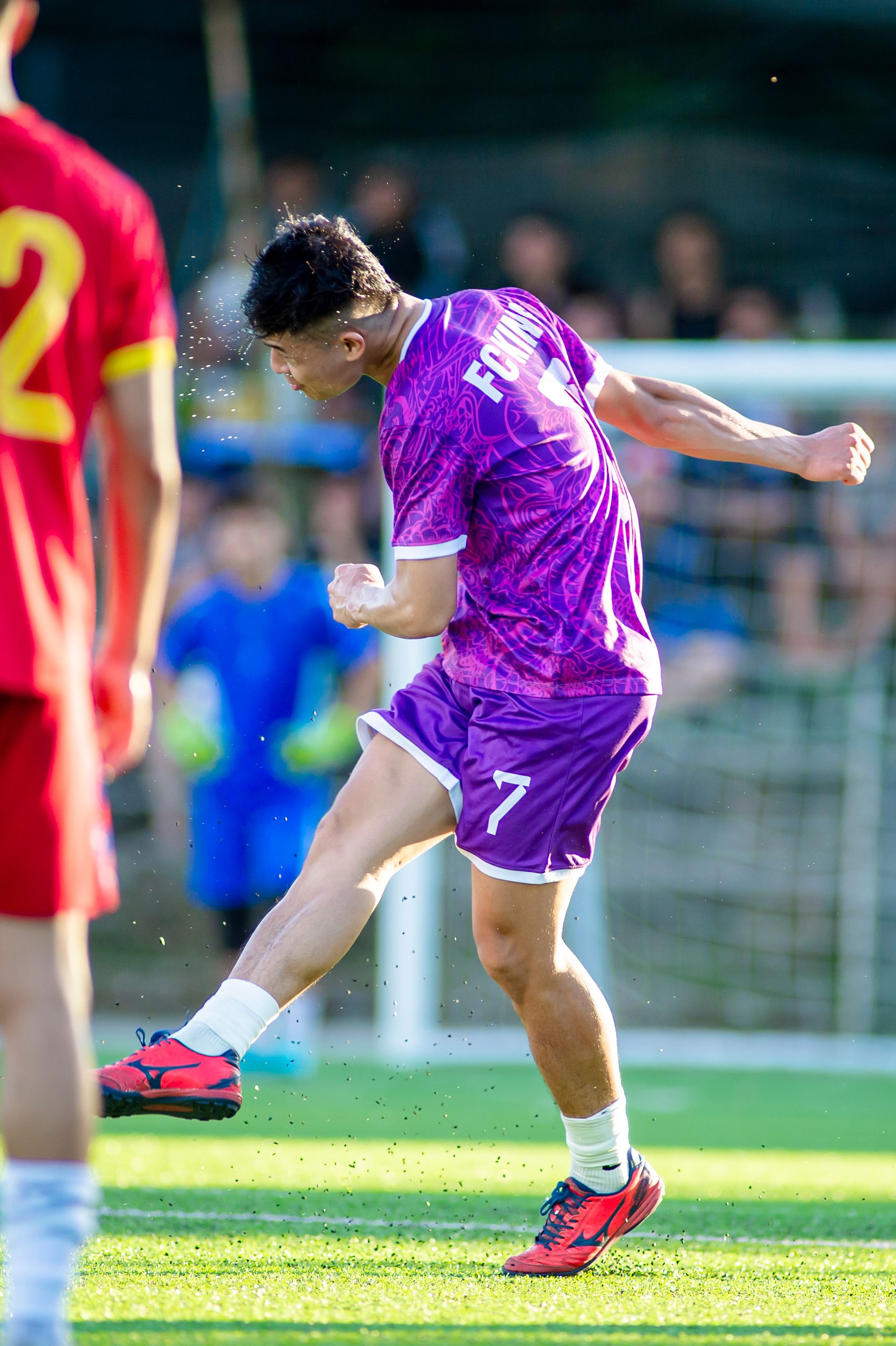 Exciting action moment of a young football player in purple uniform taking a powerful kick on a sunny day.