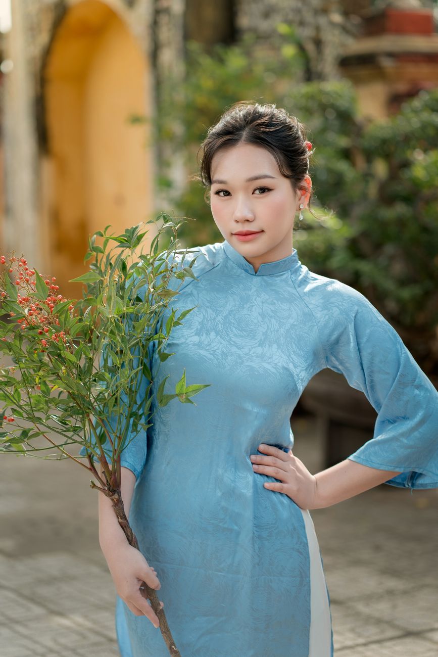 Young woman in blue áo dài holding a branch with berries in an outdoor setting.