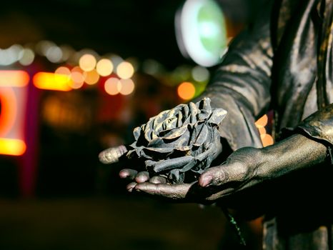Intriguing bronze sculpture of a rose held in outstretched hands, with blurred night lights in the background.
