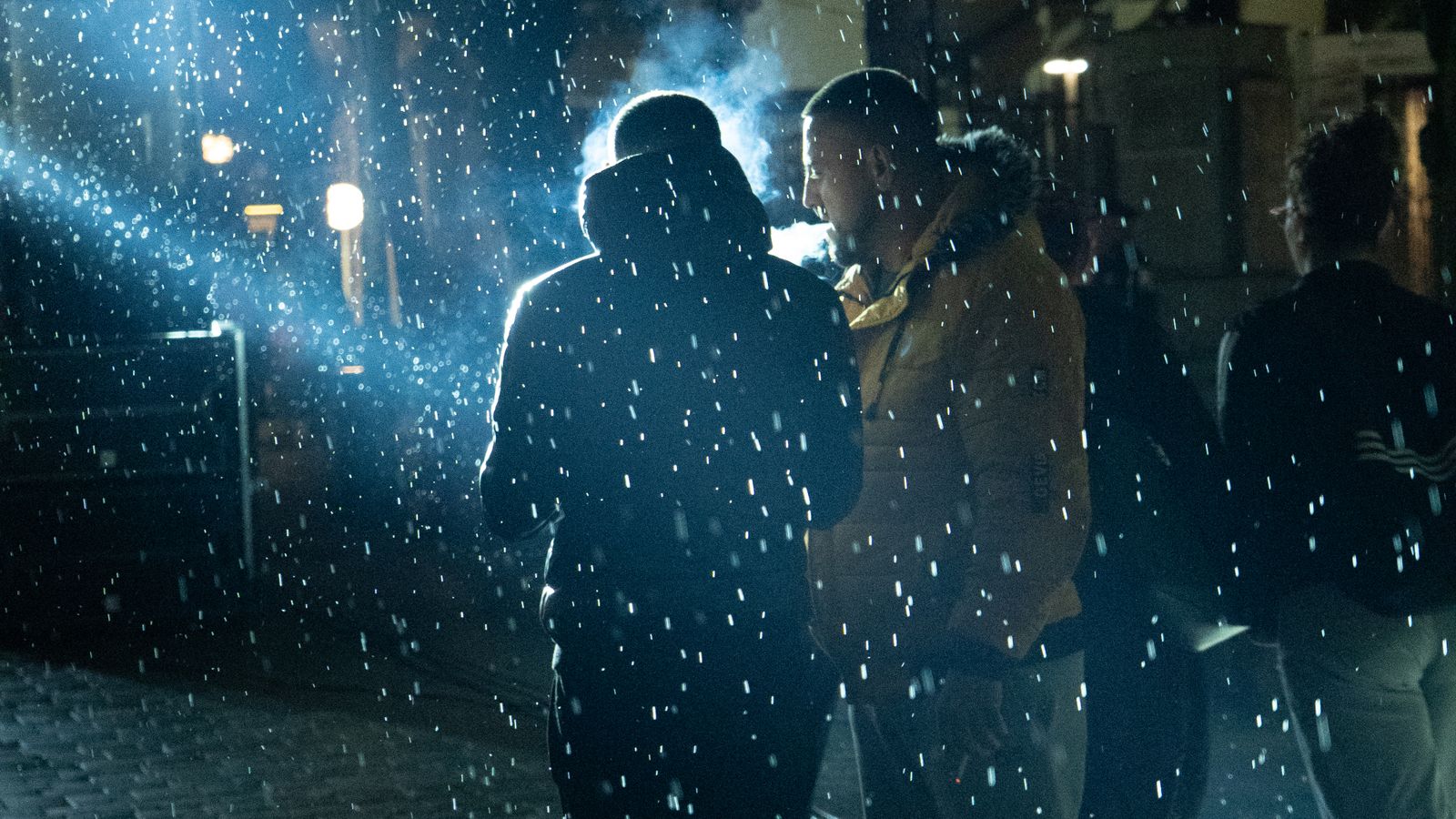 People walking under streetlights on a cobbled street as snow falls at night in Korçë, Albania