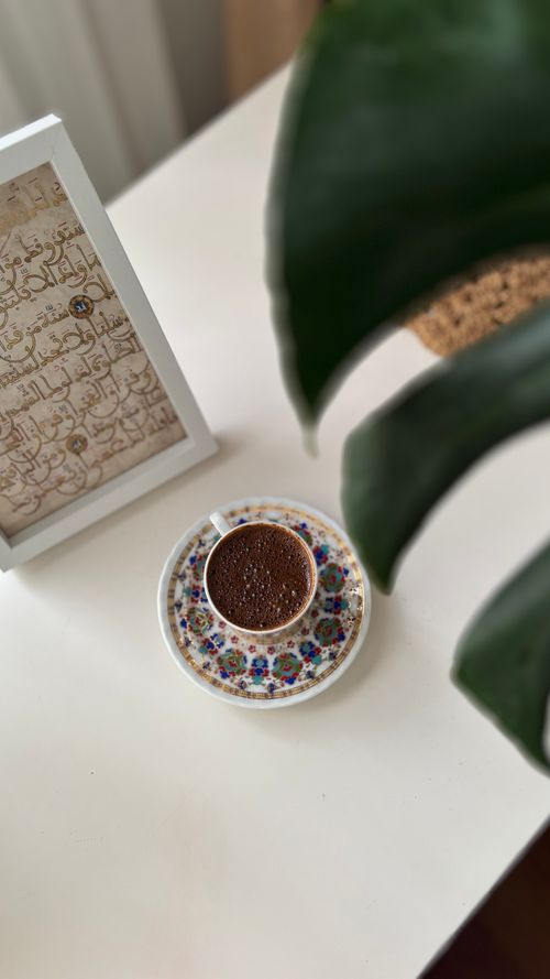 Free A cup of coffee on a white table beside framed Arabic calligraphy art and a green plant. Stock Photo