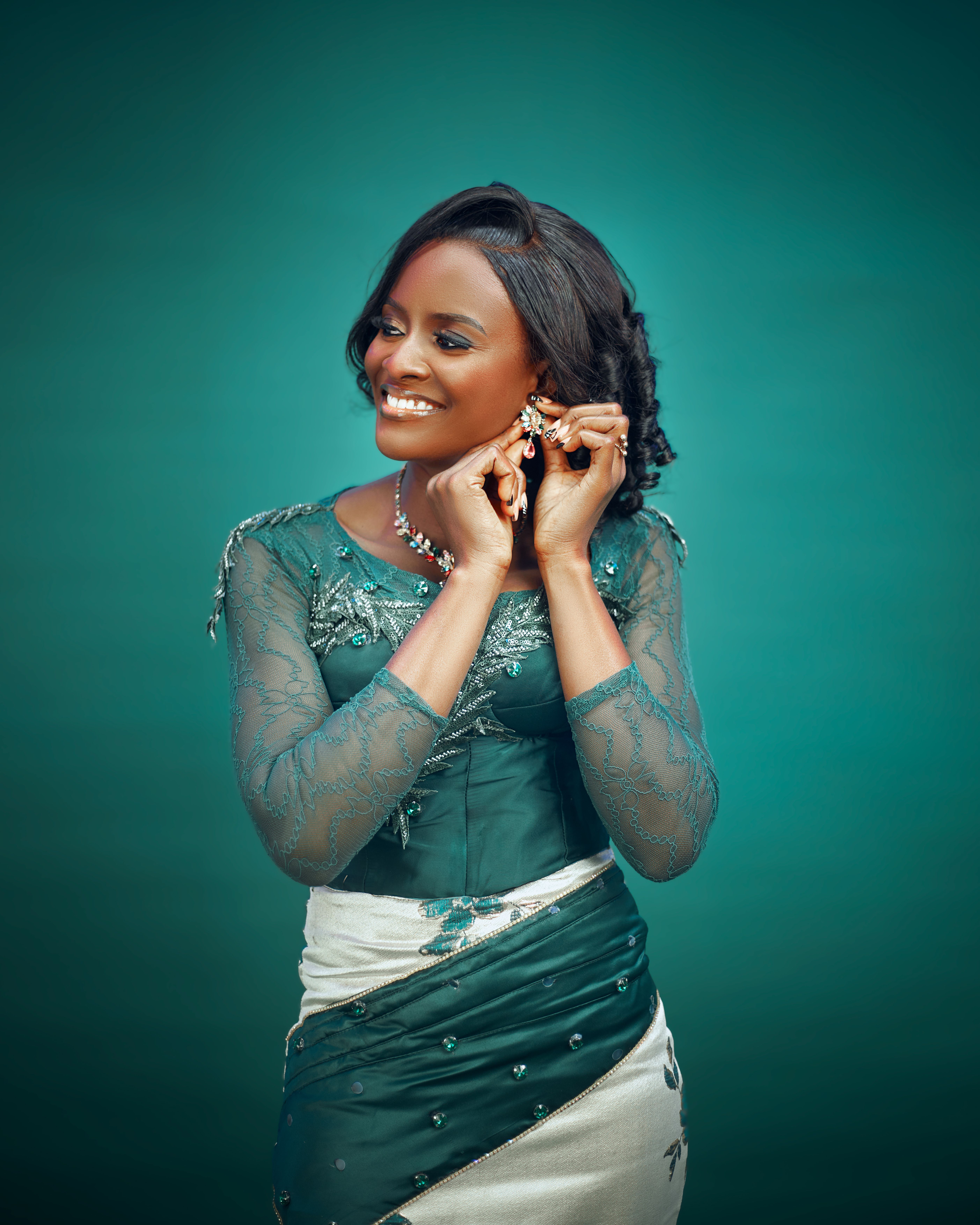 Smiling woman in elegant dress adjusting her earrings against a teal background.