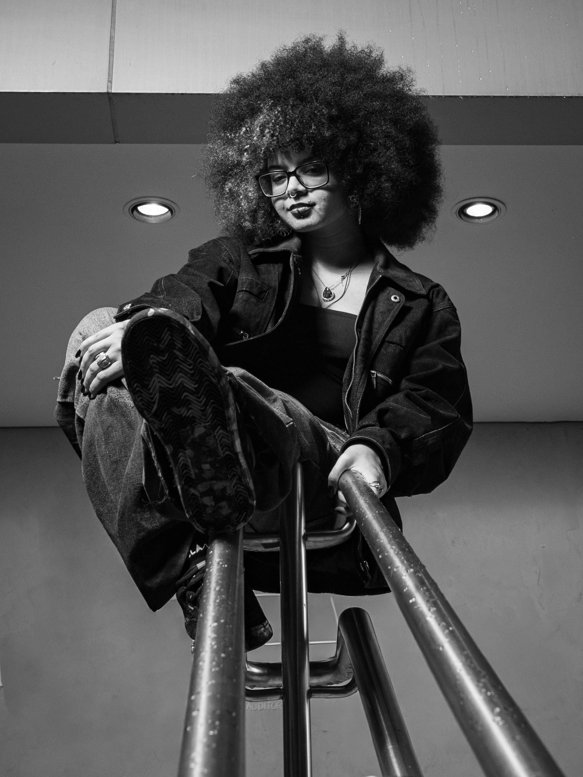 grátis Retrato em preto e branco de uma jovem de cabelos cacheados sentada em um corrimão de metal em um ambiente interno. Foto profissional